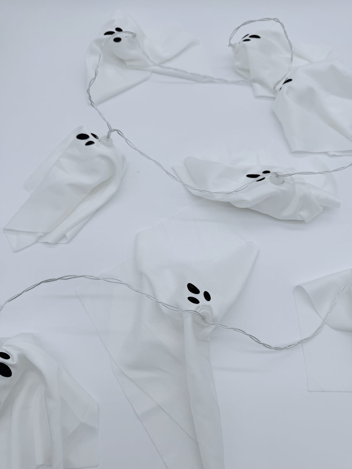 White ghost-shaped string lights with black embroidered eyes and mouths, arranged on a white background.
