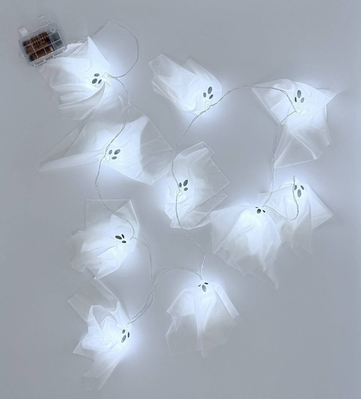 White ghost-shaped fairy string lights with small black eyes, shown illuminated against a plain light gray background. A small battery compartment box is visible in the upper left corner.
