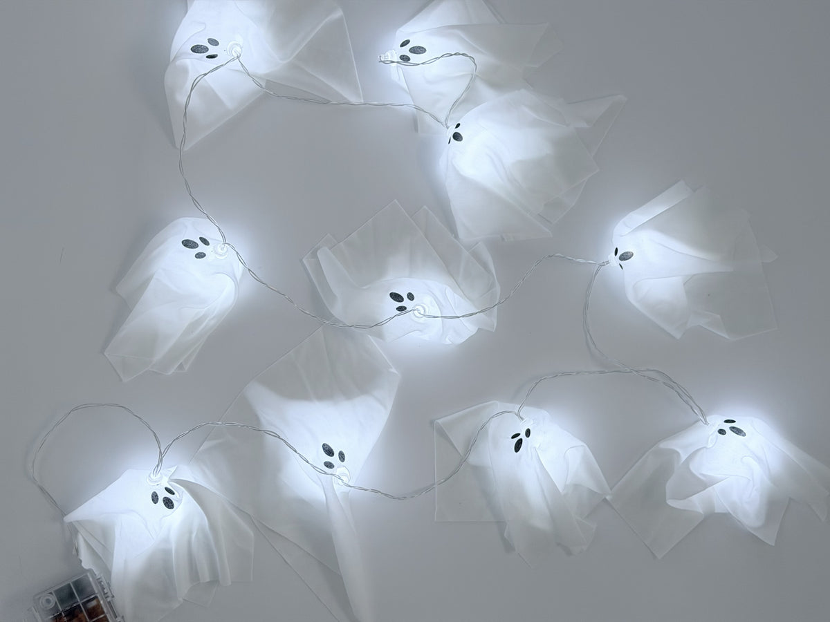 White ghost-shaped string lights with embroidered black eyes and mouths, glowing against a plain light gray background.