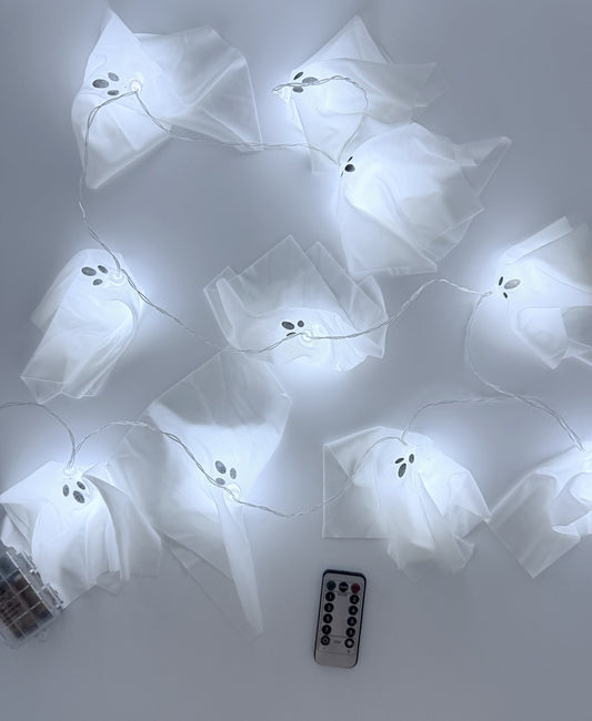 White ghost-shaped fairy string lights with a small black face detail, shown illuminated on a light gray background with a remote control and a small container of batteries visible.